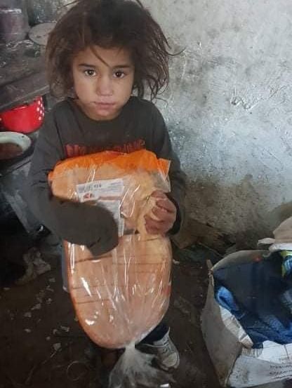 Ein junges Mädchen hält ein großes Brot, das in Plastik eingewickelt ist, und steht in einem Raum mit einem schmutzigen Boden und einer Wand im Hintergrund.