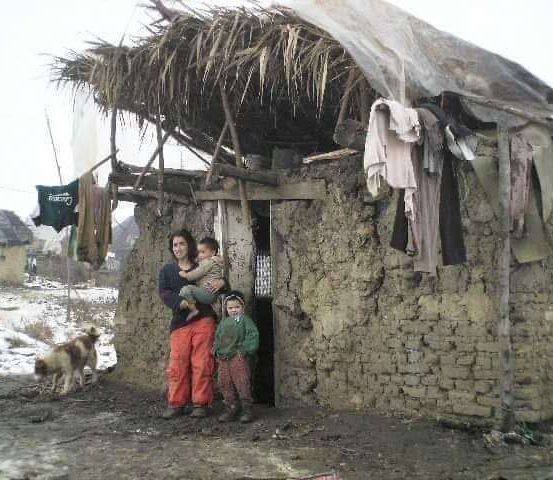 Eine Frau mit zwei Kindern steht vor einem kleinen, baufälligen Haus. Sie sind draußen, mit einem Hund in der Nähe.