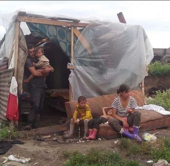 Eine vierköpfige Familie sitzt auf einer Couch in einem Behelfsunterstand mit einer Plane. Sie sind in einem Außenbereich, umgeben von Erde und Gras.