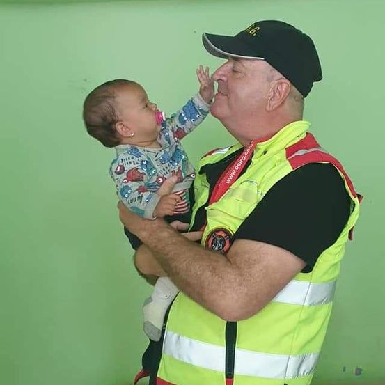 Ein Mann mit einer Warnweste hält ein Baby mit einem Schnuller. Das Baby streckt eine Hand aus.