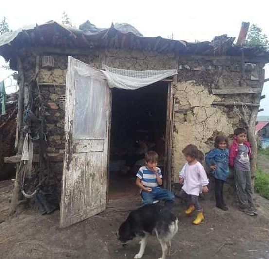 Mehrere Kinder stehen vor einem kleinen Haus mit einem Hund. Das Haus hat eine Steinmauer und eine Holztür. Ein Mann geht im Hintergrund.