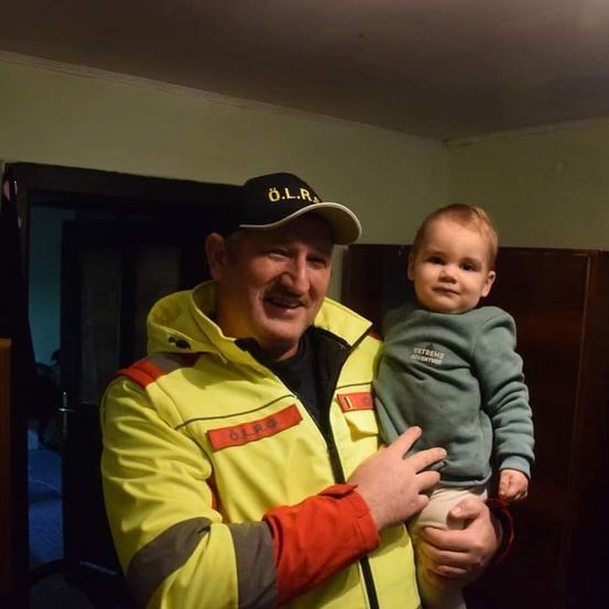 Ein Mann in einer gelben Sicherheitsjacke mit dem Wort O.L.R. hält ein lächelndes Baby. Das Baby trägt ein graues Sweatshirt mit dem Aufdruck Extreme Adventure.