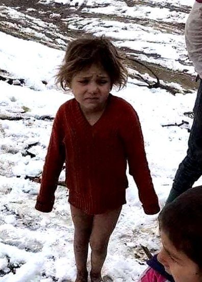 Ein junges Mädchen in einem roten Pullover steht im Schnee und sieht ernst aus. Ein anderes Kind mit einem rosa T-Shirt sieht von hinten zu.