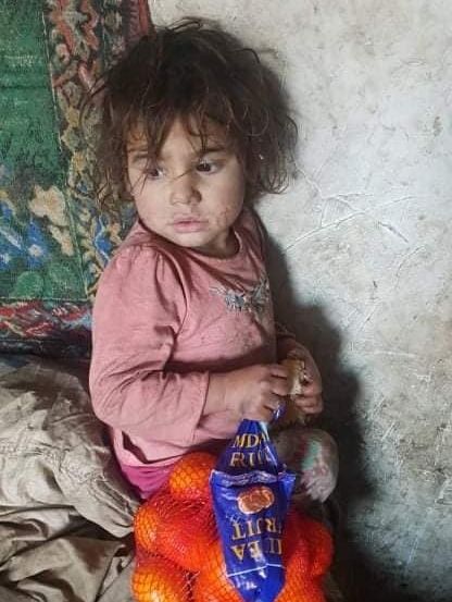 Ein kleines Kind in einem rosa T-Shirt sitzt auf einem Bett mit einer Tüte Orangen vor sich.