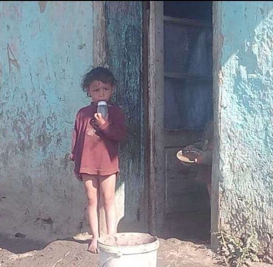 Ein Kind steht vor einem abgenutzten Haus, hält einen kleinen Behälter in der Hand, mit einem Eimer auf dem Boden und einer leicht geöffneten Tür.