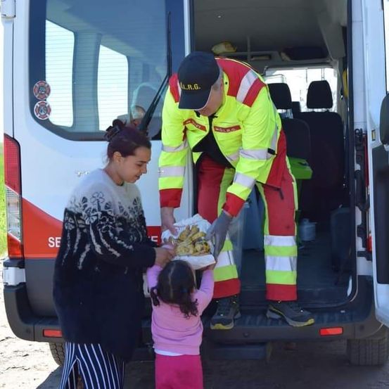Ein Mann in reflektierender Kleidung gibt einer Frau und einem Mädchen Essen durch eine offene Van-Tür.