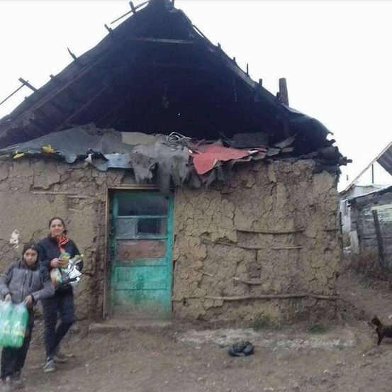 Zwei Personen, eine mit einem Schal, stehen vor einem Lehmhaus mit einem beschädigten Dach. Ein Kind hält Plastiktüten, und ein Hund ist in der Nähe.
