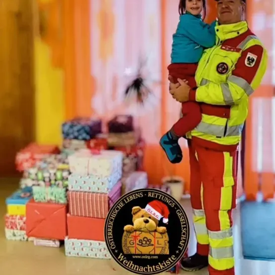 Ein Feuerwehrmann in gelber und roter Uniform hält ein Kind mit Weihnachtsmütze. Sie stehen vor einem Stapel Geschenke, möglicherweise für Weihnachten.