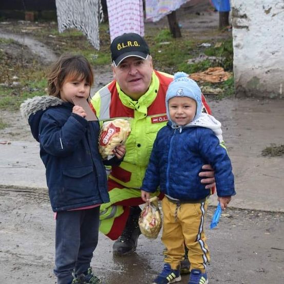 Ein Mann in einer Warnweste hält zwei Kinder, eines isst einen Snack, auf einer nassen Straße.