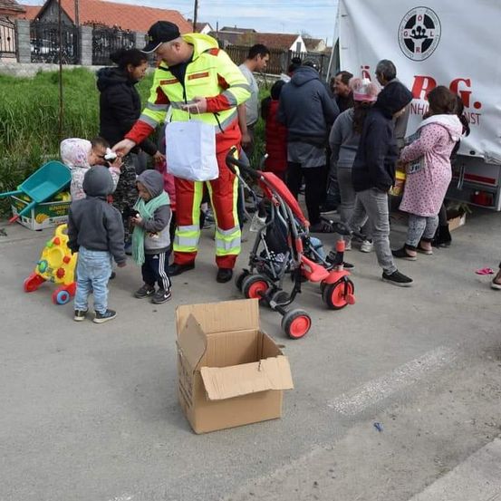Ein Mann in einer Sicherheitskleidung verteilt Gegenstände an Kinder. Eine Gruppe von Menschen, einschließlich Kindern, steht mit Kinderwagen und Spielzeug in einer Schlange. Ein Karton liegt auf dem Boden.