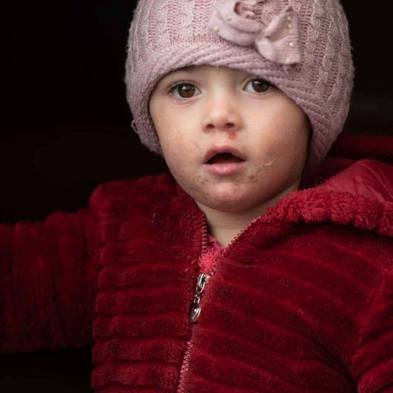 Ein Kind in einem roten Mantel mit einer rosa Mütze mit einer Blume darauf schaut nach oben. Der Mantel hat einen Reißverschluss.