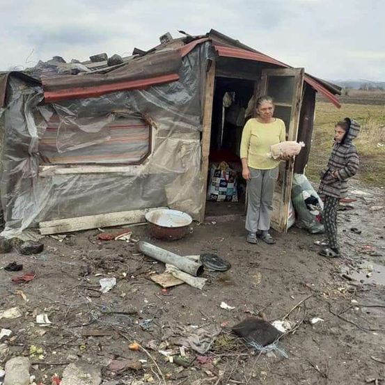 Eine Frau steht vor einer verfallenen Hütte, hält einen Plüschtier und ein Kind in der Nähe. Um die Hütte herum liegen Müll und Trümmer.