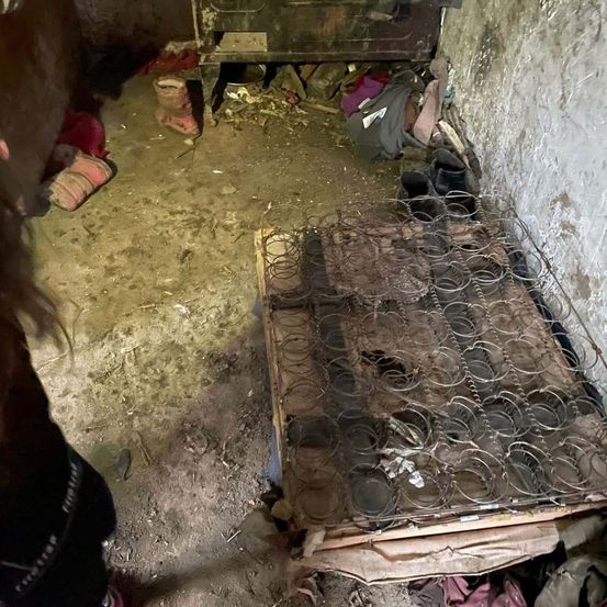 Ein verwahrlostes Zimmer mit zerbrochenem Bettgestell, verstreuten Schuhen und schmutzigem Boden. Eine Person mit Stiefeln steht in der Nähe.