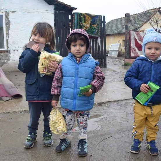 Drei junge Kinder stehen auf einer nassen Straße und halten jeweils Lebensmittelpakete in den Händen. Das Mädchen auf der linken Seite lächelt, während es isst. Der Junge in der Mitte sieht ernst aus. Der Junge auf der rechten Seite schaut nach unten. Hinter ihnen befinden sich Häuser mit Ziegelwänden.
