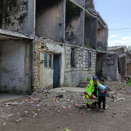 Ein Mann in einem gelben und roten Uniform hockt mit einem kleinen Jungen, der einen Beutel hält, auf einer beschädigten Straße. Dahinter befinden sich zerstörte Gebäude mit zerbrochenen Fenstern und Trümmern.
