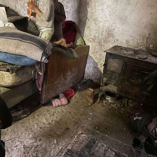 Ein Zimmer mit einem Holzbett, Decken und verstreuter Kleidung. Ein alter Ofen steht an der Wand und der Boden ist unordentlich.