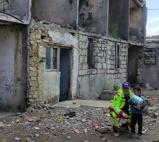 Ein Mann in einer grünen Uniform hält ein Kind in seinen Armen und steht vor einem beschädigten Gebäude.