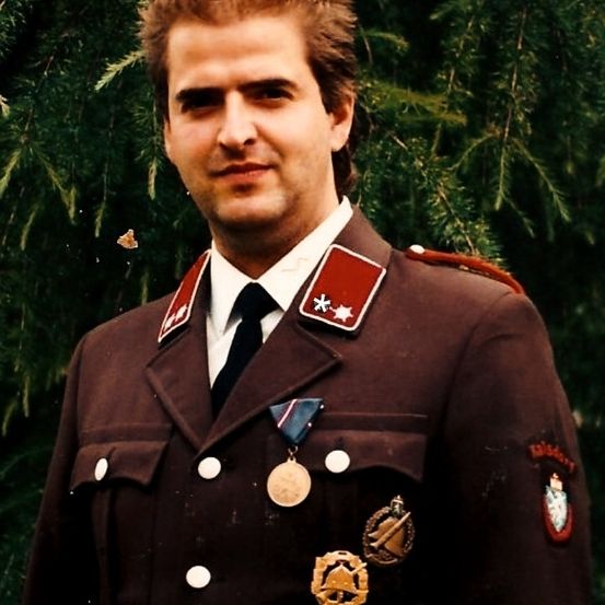 Ein Mann in Uniform steht vor einem Baum, trägt Medaillen und einen roten Aufnäher am Ärmel.