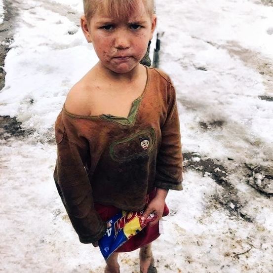 Ein junger Junge steht im Schnee, trägt ein zerrissenes Shirt mit Dreck darauf und hält einen Snack in der Hand.