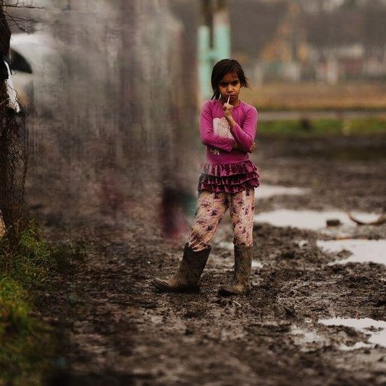Ein kleines Mädchen steht auf schlammigem Gelände, trägt ein rosa Shirt und Stiefel und hält eine Zigarette.