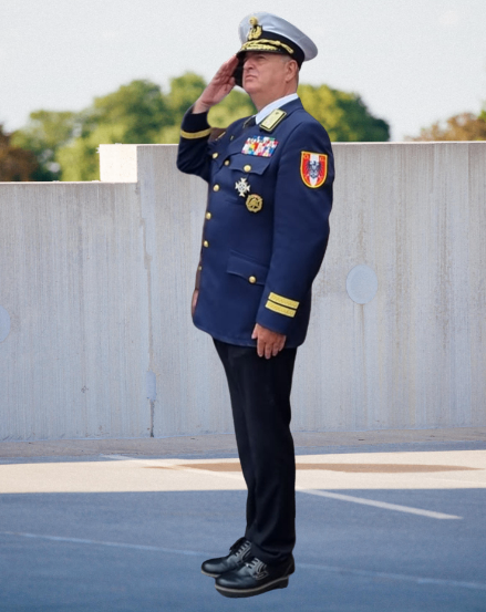 Ein Mann in Marineuniform steht und salutiert. Er trägt einen Hut mit Goldbesatz und mehrere Medaillen auf der Brust.