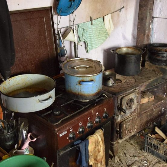 Eine alte Küche mit einem rostigen Herd und Töpfen darauf. Ein grünes Handtuch hängt an der Wand und ein blauer Teller ist auf dem Gestell.