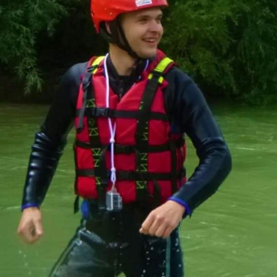 Ein Mann mit rotem Helm und Schwimmweste steht lächelnd im grünen Wasser.