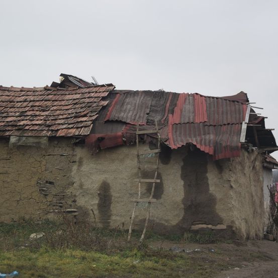Ein altes Haus mit einem beschädigten Dach, bedeckt mit roten Blechen, lehnt sich gegen eine Wand. Eine hölzerne Leiter ist an der Seite platziert.