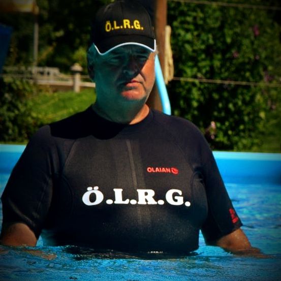 Ein Mann in einem schwarzen Neoprenanzug mit den Buchstaben 'O.L.R.G.' steht im Pool. Er trägt einen Hut mit den gleichen Buchstaben.