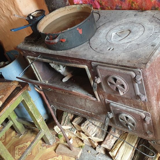 Ein alter Ofen mit einem Topf darauf steht in einem Raum mit einem Holzstuhl und einem Holzstapel. Eine Katze sitzt im Inneren des Ofens.