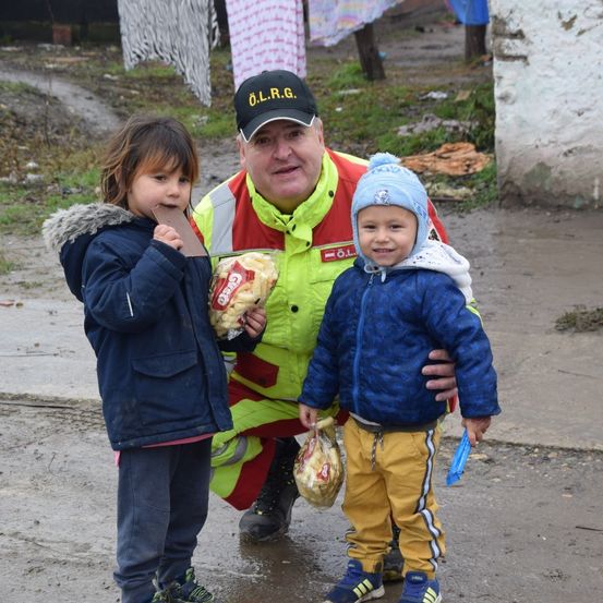 Ein Feuerwehrmann in einer reflektierenden Weste teilt einen Snack mit zwei Kindern. Die Kinder halten Taschen und eines isst eine Schokoladentafel. Der Hintergrund ist unscharf.