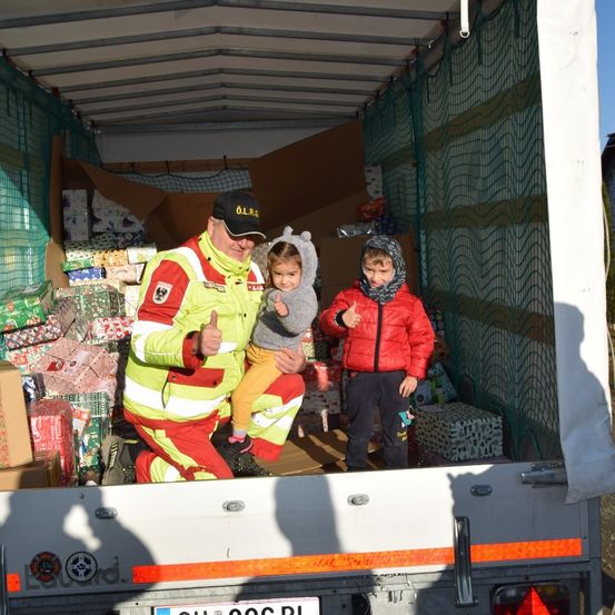 Ein Feuerwehrmann in gelber und roter Uniform hält ein Kind auf dem Schoß, während er im hinteren Teil eines mit Geschenken gefüllten Lkw steht.