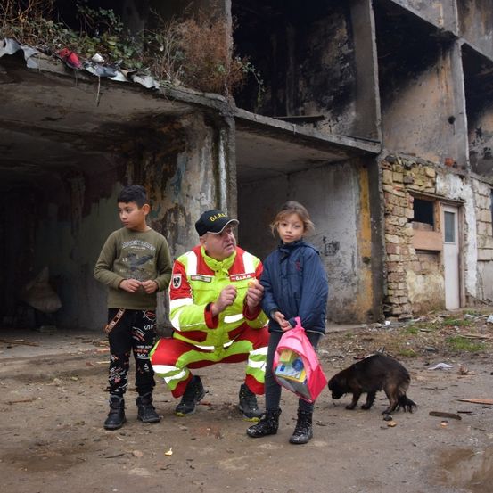 Ein Mann in einem Schutzanzug kniet vor zwei Kindern und einem Hund vor einem beschädigten Gebäude.