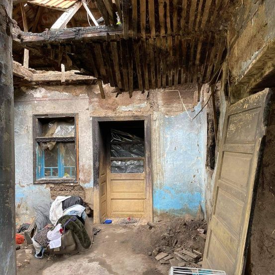 Ein verlassener Raum mit beschädigter Decke, einer Holztür und einem Fenster. Der Boden ist mit Schutt bedeckt, in der Nähe befindet sich ein Haufen Kleidung.