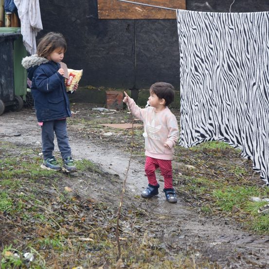 Zwei kleine Kinder stehen in einem schlammigen Bereich mit Müll um sie herum. Eines hält eine Tüte Chips, und das andere zeigt auf etwas. Ein Zebra-Mustertuch hängt rechts.