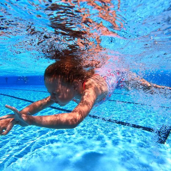 Ein Mädchen schwimmt unter Wasser in einem Pool. Sie trägt einen Badeanzug und hat ihre Hände leicht angehoben. Das Wasser ist klar und sie ist von Wasserspritzern umgeben.