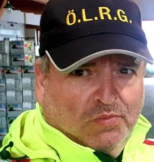 Ein Mann in einer reflektierenden Weste und Mütze mit gelben Buchstaben, die O.L.R.G. stehen, posiert vor einem Laden.