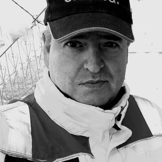 Ein Mann mit ernster Miene trägt eine schwarze Kappe mit weißen Buchstaben und eine weiße Jacke mit reflektierenden Streifen. Er steht vor einem Zaun.