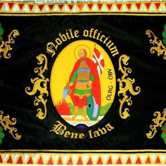 Ein schwarzer Banner mit goldenen Designs zeigt ein zentrales Emblem. Es enthält einen Ritter mit Schwert und Schild, mit lateinischen Inschriften darum herum. Die Ecken des Banners haben dekorative Elemente.