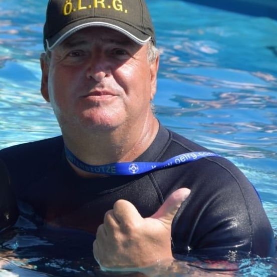 Ein Mann mit grauem Haar in einem schwarzen Neoprenanzug mit O.L.R.G.-Kappe ist im Pool und gibt einen Daumen hoch.