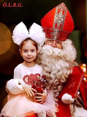 Ein junges Mädchen in einem rosa Kleid hält ein Geschenk und lächelt, posiert mit dem Weihnachtsmann, der einen roten Hut und ein rotes Buch trägt.