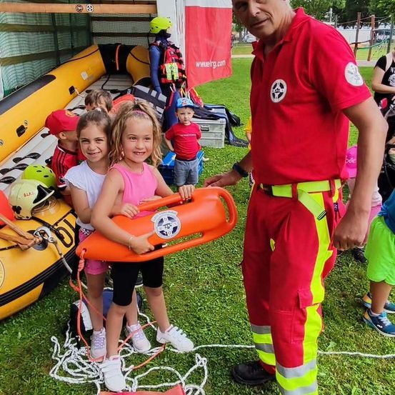Ein Mann in einem roten Uniform steht mit zwei Mädchen, die einen orangefarbenen Rettungsring halten, mit Kindern und aufblasbaren Booten im Hintergrund.