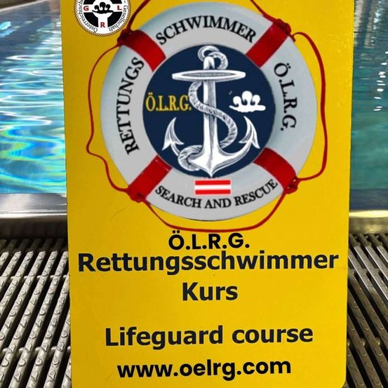 Ein gelbes Schild bewirbt einen Rettungsschwimmer-Kurs. Es zeigt einen Rettungsring mit einer Flagge und einem Anker, mit der Aufschrift 'Suche und Rettung'. Darunter steht 'O.L.R.G. Rettungschwimmer Kurs' und 'Lifeguard course'. Es endet mit der Website www.oelrg.com.