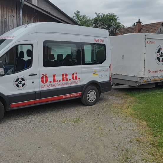 Ein weißer Van mit rotem Schriftzug steht auf Schotter neben einem Holzgebäude. Der Van hat rote Sitze, und der Anhänger dahinter ist weiß.