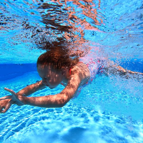 Ein junges Mädchen schwimmt im Pool mit dem Brustschwimmen. Sie hat einen Badeanzug an und ist vollständig untergetaucht. Ihre Haare sind nass.