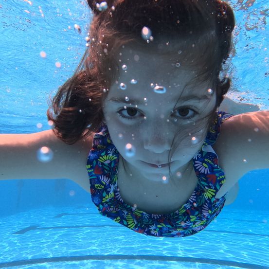 Ein junges Mädchen mit braunen Haaren schwimmt unter Wasser in einem Pool. Sie trägt ein farbenfrohes Badeanzug mit Blumen. Blasen umgeben sie, und ihr Gesicht ist über dem Wasser sichtbar.