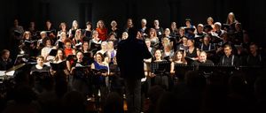 Ein Chor tritt auf der Bühne auf, mit Notenblättern vor ihnen, beleuchtet von einem Scheinwerfer. Dahinter steht ein Dirigent, und davor schaut ein Publikum zu.