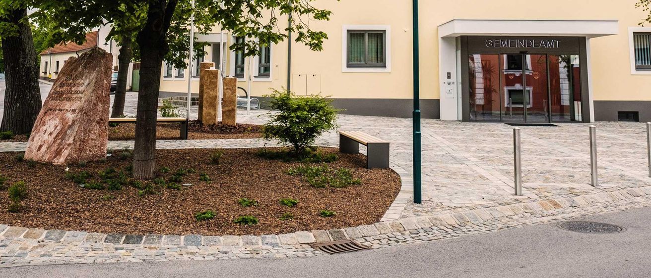 Ein Außenbereich mit einem Baum, Bänken, einer Straßenlaterne und einem Gebäude mit rotem Dach und Fenstern, mit einem Schild, das 'Hauptstraße' steht.