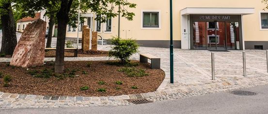 Ein Außenbereich mit einem Baum, Bänken, einer Straßenlaterne und einem Gebäude mit rotem Dach und Fenstern, mit einem Schild, das 'Hauptstraße' steht.