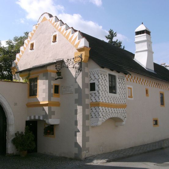 Ein kleines traditionelles Haus mit schwarzem Dach und gelber Einfassung, mit kunstvollen Schnitzereien an der Fassade. Eine Lampe hängt über dem Eingang an der Wand.